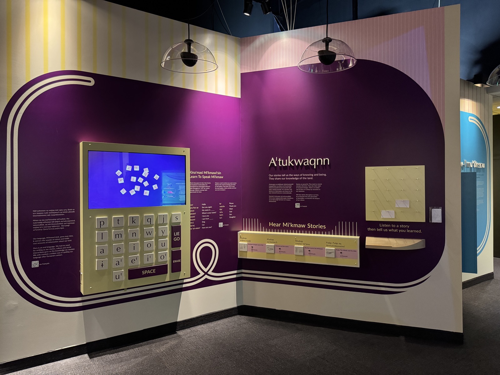 Two walls of the Pjila'si gallery: deep purple panels with looping cream line graphics, a brass-framed interactive Mi'kmaw keyboard and screen, and an adjacent &ldquo;A'tukwaqnn&rdquo; storytelling wall with a Hear Mi'kmaw Stories listening bar.